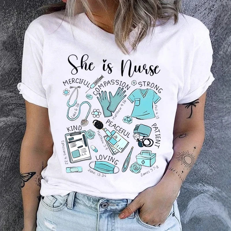 A woman wearing a white graphic t-shirt with a black and teal print that reads 'She is Nurse' surrounded by medical icons and words like compassion, kindness, patience, and loyalty. She is also wearing light-wash denim jeans, standing against a textured gray wall.