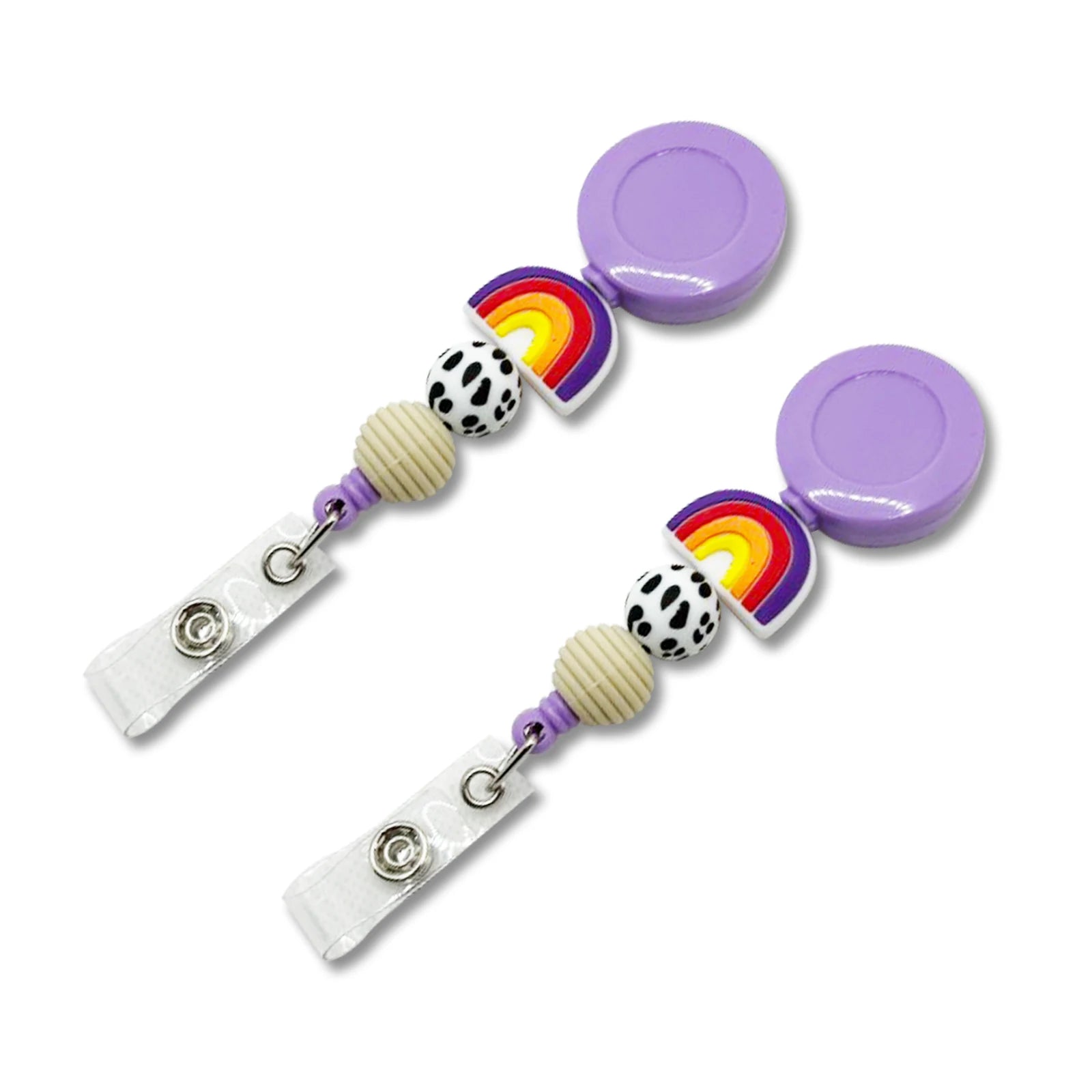 Two matching retractable badge reel clips on a white background. Each clip has a purple round reeling cap, a small rainbow charm, a black-and-white spotted bead, a cream ribbed bead, and a clear plastic clip with a metal snap.