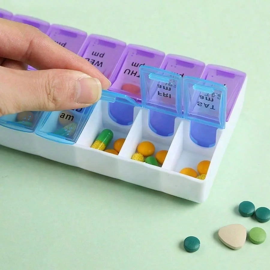 A weekly pill organizer tray with purple and blue compartments, shown with a hand placing a pill into one of the open slots. Several loose pills are scattered on a pale mint green surface.