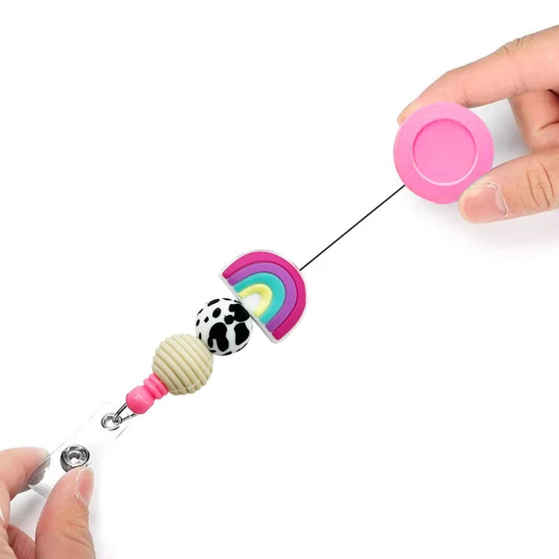 A retractable baby clip with a colorful rainbow design and cow-print bead, held by hands on a white background with descriptive text.