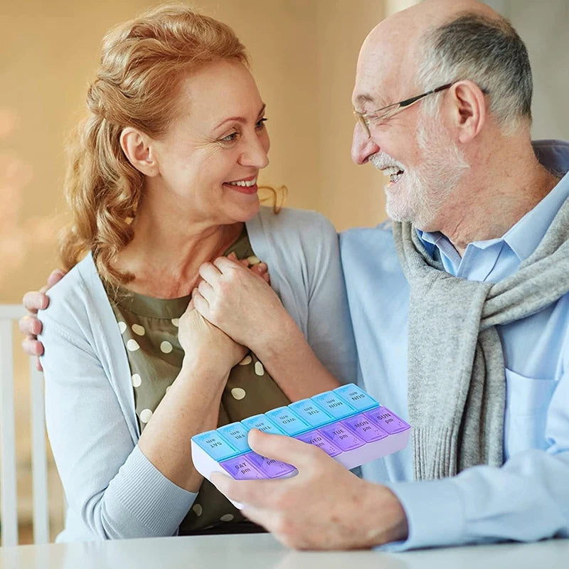 An older man and woman are smiling and hugging indoors while the man holds a pastel-colored weekly pill organizer with blue, green, and purple compartments.